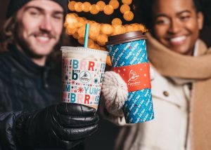 Two people holding Caribou Coffee Holiday cups, one cold cup, one hot. The cups say "Brrrum Pum Pum Yum" and "Sleigh All Day" as part of a pattern.